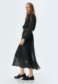 Zwarte geplisseerde jurk met polkadots, lange doorzichtige mouwen, hoge halslijn, tailleband en gelaagde zoom. Draag het met zwarte loafers.