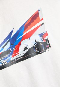 Balta marškinėliai su spalvingu grafikų dizainu, vaizduojančiu lenktyninį automobilį su mėlynais, raudonais ir baltais trikampiais, taip pat aiškiai matomomis padangų detalėmis.