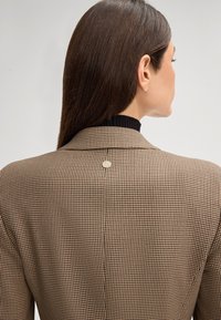 Blazer a quadri marrone con una superficie liscia, rever a punta e un piccolo logo argentato sul retro.