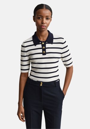 Kvinde iført en hvid kortærmet strikket polo med marineblå vandrette striber og marineblå bukser med guldknapdetaljer.