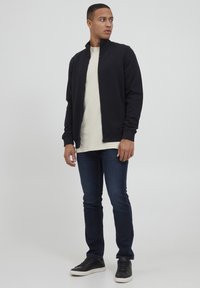 Svart zip-up jakke over en lys farget t-skjorte, kombinert med mørkeblå jeans og svarte sneakers, har et minimalistisk design med rillekantet krage.