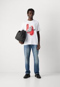 Junger Mann hält eine schwarze Tasche, trägt ein weißes T-Shirt mit roten Boxhandschuhen und einem Boxer-Grafik, blaue Jeans und schwarze Turnschuhe, steht vor einem weißen Hintergrund.