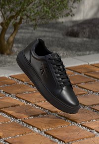 Fekete bőrsneaker fűzőkkel és Calvin Klein Jeans logóval, lebeg a fából készült négyzet alakú csempék felett, kavicsos hézagokkal a szabadban.