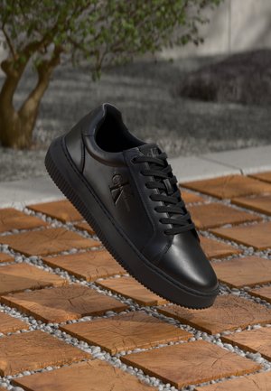 Zwarte leren sneaker met veters en het Calvin Klein Jeans-logo, zwevend boven houten vierkante tegels met grindgaten buitenshuis.