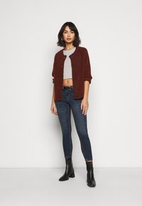 Cardigã castanho de malha com mangas curtas, frente com botões, combinado com um top crop cinzento e jeans skinny escuros, finalizado com botas de salto pretas.