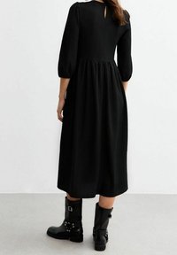 Musta midi-mekko puffihihnoilla, teksturoidusta kankaasta ja takana avokeyhole-yksityiskohta, yhdistettynä mustiin nilkkasaappaisiin, joissa on solkia.