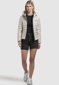 Lichtgrijze pufferjas met horizontale padding, een zilvergekleurde capuchon en een ritssluiting aan de voorkant. Draagt bij zwarte shorts en lichte sneakers.