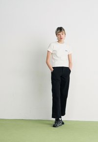 T-shirt bianco a maniche corte aderente con un piccolo logo, abbinato a pantaloni neri a gamba larga e sneakers nere, su un pavimento verde.