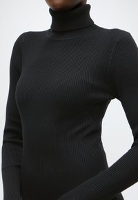 Pull à col roulé noir côtelé avec manches longues, présentant une coupe ajustée et un tissu texturé qui met en valeur la silhouette.