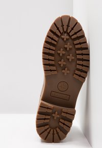 Semelle d'une botte Timberland marron avec des crampons profonds, des rainures croisées et droites, et le logo Timberland embossé près du talon.