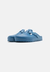 Zuecos de plástico azul con forma redondeada, parte trasera abierta y una correa ajustable en la parte superior. Suela texturizada para un mejor agarre.