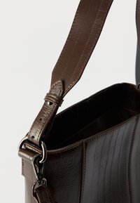 Borsa a mano in pelle marrone con superficie testurizzata, dotata di una tracolla larga e accessori metallici per la regolazione. L'apertura superiore lascia intravedere i dettagli interni.