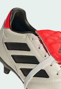 Botín de fútbol con un upper de cuero blanco, acentos negros y una lengüeta acolchada de color rojo brillante. Presenta un cierre con cordones y una superficie texturizada.