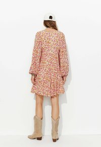 Robe à imprimé floral avec de longues manches bouffantes, des poignets élastiques et un ourlet en niveaux. L'arrière-plan est clair, avec un mannequin portant des bottes de cowboy beiges et une casquette.