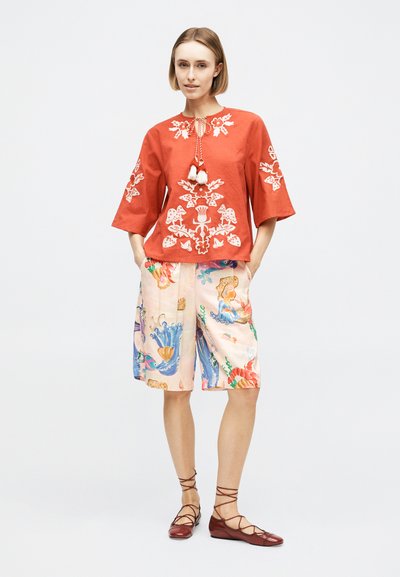 Femme portant un chemisier rouge brodé avec des pompons, un short coloré à motifs jusqu'aux genoux, et des chaussures plates marron à lacets, debout les mains dans les poches.