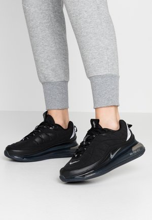 Fødder iført sorte Nike Air Max 720 sneakers med synlige luftsåler, kombineret med lysegrå opsmøgede joggingbukser mod en ensfarvet baggrund.