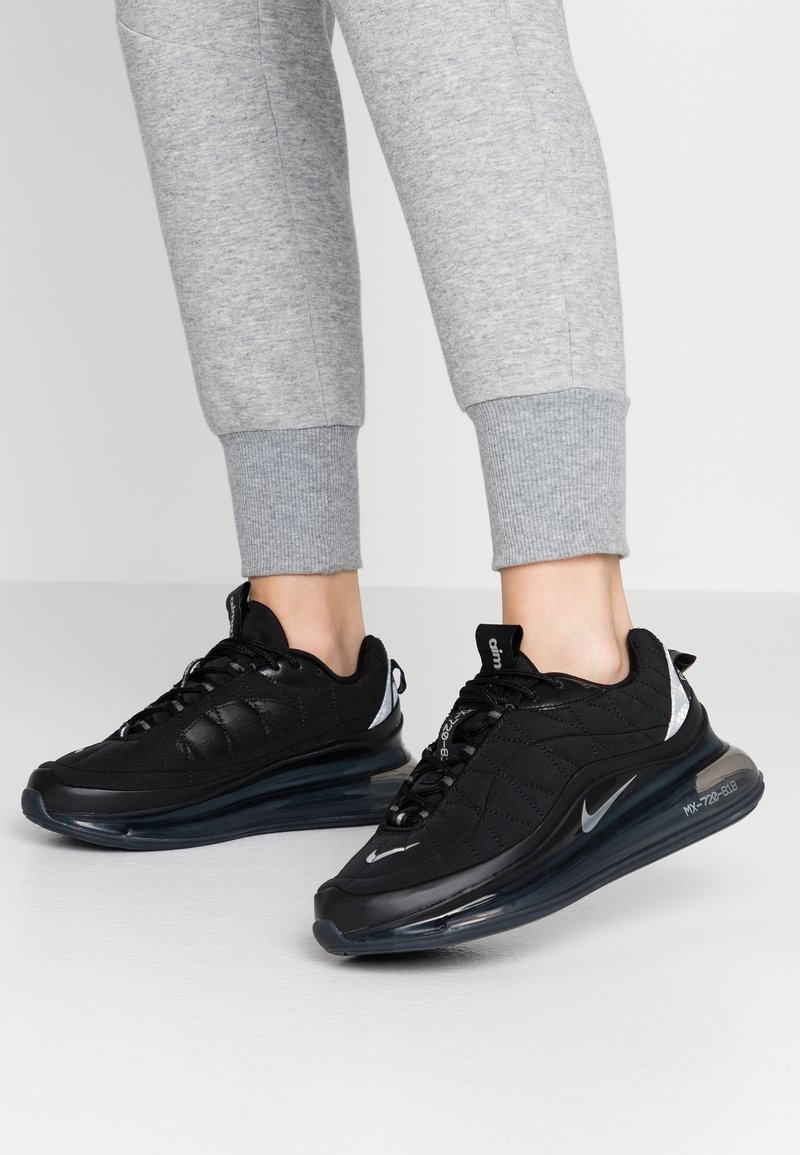 Pieds chaussés de baskets noires Nike Air Max 720 avec semelles à air visibles, associées à un pantalon de jogging gris clair retroussé sur un fond uni.