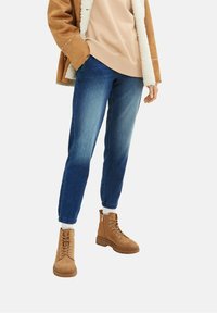 Personne portant un jean bleu ajusté, des bottines à lacets couleur camel, un sweat-shirt beige et une veste doublée en peau retournée camel, debout avec une main dans la poche.