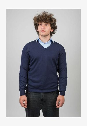 Marinblaue V-Ausschnitt-Strickjacke aus weichem Stoff, kombiniert mit einem hellblauen Hemd mit Kragen. Der Mann steht mit einem neutralen Ausdruck vor einem grauen Hintergrund.