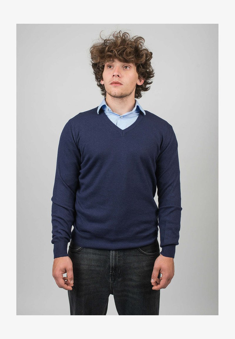 Námorně modrý svetr s výstřihem do V z jemné pleteniny, v kombinaci se světle modrou košilí s límečkem. Muž stojí s neutrálním výrazem na šedém pozadí.