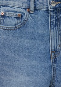 Primer plano de unos jeans de mezclilla azul claro que muestran el bolsillo frontal, el lazo del cinturón, el botón y los detalles de costura naranja.