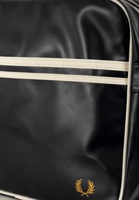 Borsa in pelle nera con due strisce beige, caratterizzata da un emblema dorato nella parte inferiore. Texture liscia, forma rettangolare e chiusura con zip robusta.