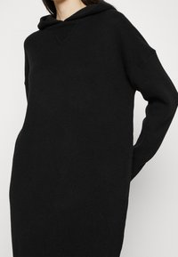 Abito maglione con cappuccio nero, realizzato in morbido tessuto a maglia. Presenta una vestibilità comoda, maniche lunghe e un piccolo dettaglio sul collo a V in texture.