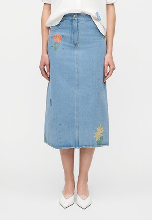 Średniej długości jasnoniebieska spódnica dżinsowa z haftowanymi pomarańczowymi i żółtymi kwiatami, noszona z białymi szpilkami o spiczastych noskach.