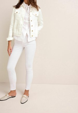 Woman wearing white denim jacket, white button-up shirt, skinny white jeans, and cream-colored slip-on loafers against beige background.