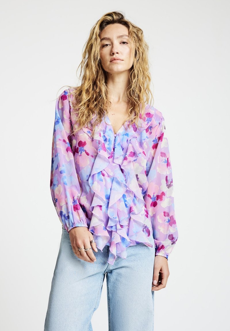 Transparente Blouson-Oberteil mit Rüschen vorne in pastellrosa, lila und blauem Blumenmuster. Lange Ärmel mit elastischen Bündchen, kombiniert mit hellen Jeans.
