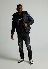 Must pufferjakk karvase äärega kapuutsiga, mille juurde on kombineeritud must graafilise mustriga dressipluus ja kulunud tumedad teksapüksid, viimistletud mustade ja valgete tennistega.