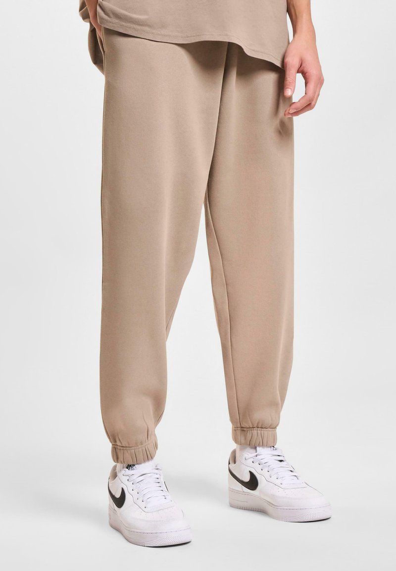 Beige Jogginghosen mit elastischen Bündchen, aus weichem Material, im lässigen Schnitt. Getragen mit weißen Sneakern mit schwarzen Akzenten.