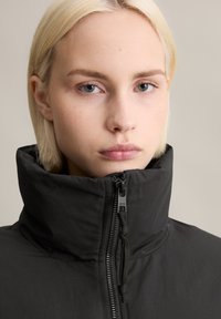 Schwarze Daunenjacke mit hohem Kragen und Reißverschlussdetails. Der Stoff wirkt glatt und matt und verfügt über einen Kordelzug am Kragen.