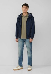 Marinefarbene Zip-Jacke über einem olivgrünen Poloshirt, hellblaue Jeans und beige Sneaker mit weißen und sandfarbenen Akzenten, stehend vor einem neutralen Hintergrund.