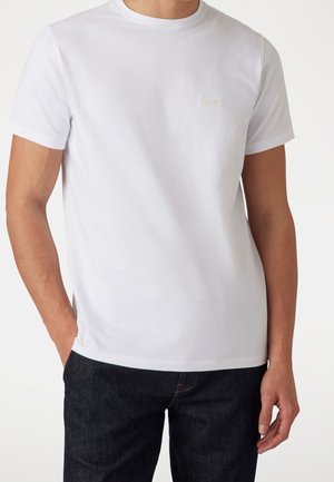 Man draagt een effen wit T-shirt met korte mouwen en donkere spijkerbroek, met één hand in de zak, getoond van schouders tot heupen.