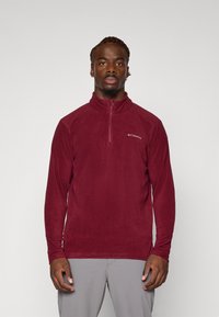 Rød fleece pullover med halv-lynlås krage, lange ermer, og en subtil logo detalj på venstre bryst. Myk tekstur, lett design.