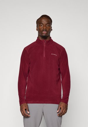 Rød fleecepullover med halv-zip krave, lange ærmer og en diskret logo på venstre bryst. Blød tekstur, let design.