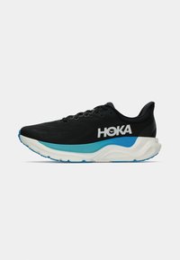 Sapatilha de corrida HOKA preta com sola almofadada branca e azul, logo lateral e design de atacadores, exibida sobre um fundo cinza liso.