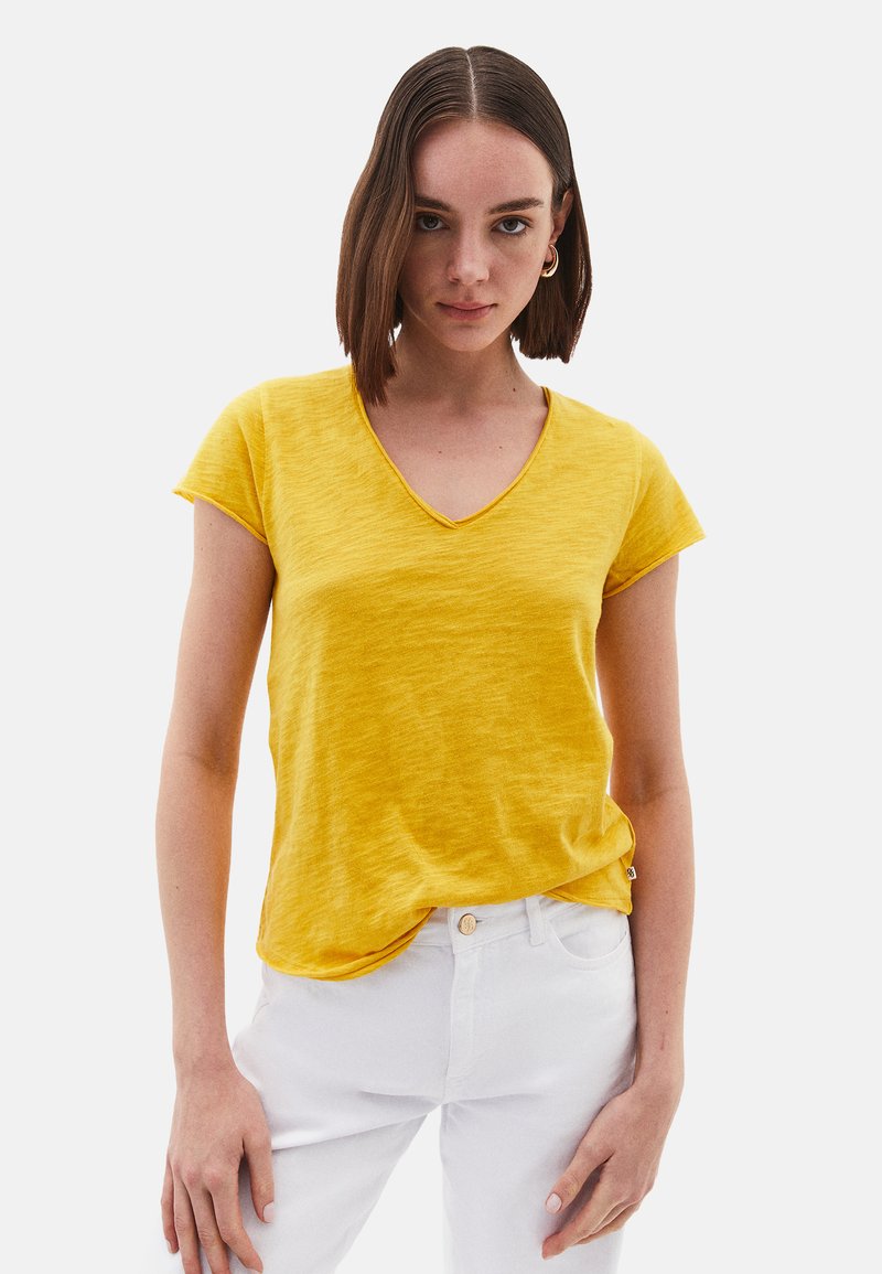 T-shirt jaune à manches courtes et col en V en tissu texturé, associé à un pantalon blanc. Présente une coupe légèrement décontractée et un ourlet incurvé.