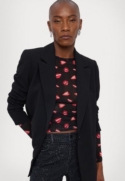 Blazer negro entallado sobre una blusa transparente con patrón de labios rojos, combinado con jeans oscuros con adornos de pedrería.