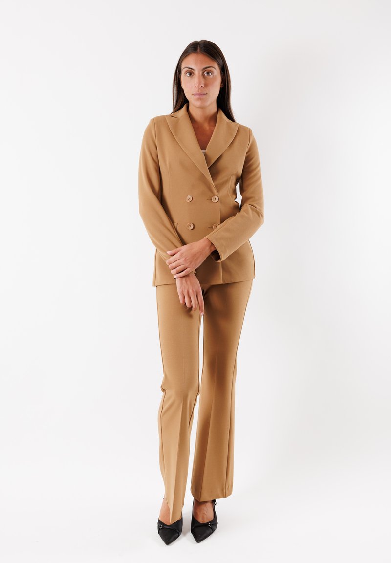Blazer doppiopetto e pantaloni svasati abbinati in tessuto beige. Collo a risvolto, due bottoni, silhouette aderente, abbinato a tacchi neri.