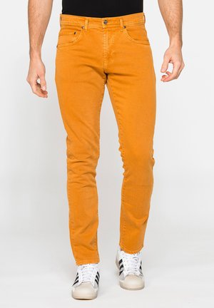 Uomo che indossa pantaloni arancioni slim-fit e sneakers bianche con strisce nere, in piedi davanti a uno sfondo chiaro e uniforme.