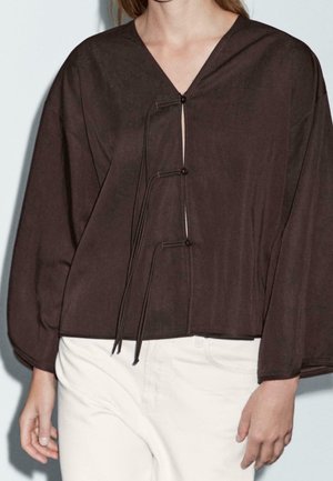 Femme portant un haut marron ample à manches longues avec trois liens à l'avant et un pantalon blanc cassé, visible du cou aux hanches sur un fond uni.