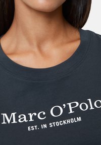 Woman's neck and upper chest wearing a dark blue shirt with "Marc O’Polo EST. IN STOCKHOLM" printed in white on the front.