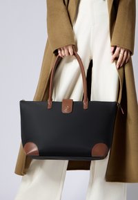 Černá taška přes rameno s hnědými koženými uchy a akcenty, která se vyznačuje elegantním designem, zipovým uzávěrem a zaoblenými rohy. Viditelné detaily z texturované látky.