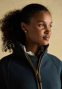 Námornícky fleecový kabát s vysokým golierom a hnedým zipsom. Má rebrovaný výstrih a pod ním bielu košeľu s krajkovým lemovaním. Zlato farbené náušnice.