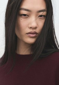 Gros plan d'une femme aux longs cheveux noirs et raides, portant un pull bordeaux. Son expression est neutre, avec des traits doux et un teint lisse.