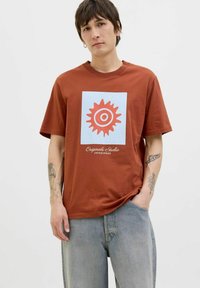 Brun bomulds t-shirt med et blåt kvadratisk grafik med et rødt solmotiv og trykt tekst nedenfor. Tekstureret materiale, korte ærmer, afslappet pasform.