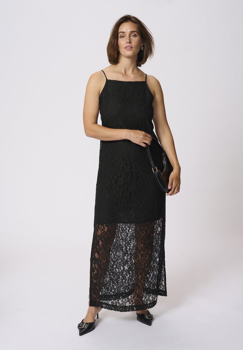 Abito maxi in pizzo nero con spalline sottili; texture intricata, design a strati con una parte inferiore trasparente. Abbinato a scarpe nere con tacco e una borsa.