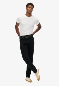 T-shirt en coton blanc, pantalon en denim noir, ceinture noire et baskets beiges. Design simple, coupe ajustée, avec des détails de couture visibles.